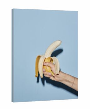 Caucasian young adult woman hand holding partially peeled banana against blue background, fingers gripping fruit firmly, nails painted, wrist adorned with thin bracelet, casting shadow