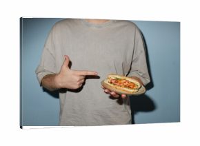 Caucasian young adult man holding plate with sandwich and pointing at food, standing against plain blue background, only upper body and hands visible, face not shown
