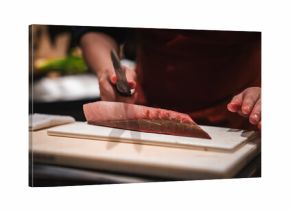 A chef's hands slice a thick tuna fillet with a yanagiba on a white board at a Japanese hotel sushi counter, dim focused lighting and warm tones emphasize craft.