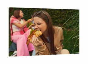 Young woman biting burger with passion while friends laugh nearby. Concept of authentic emotion, youth lifestyle, food realism for street-food branding, social content, and lifestyle marketing.