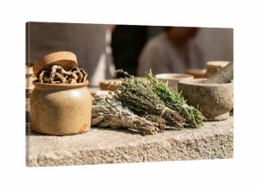Dried herbs and mortar sit on a rustic stone market table with ceramic jars and woven baskets suggesting traditional apothecary or culinary preparation. Blurred background provides copyspace for text 