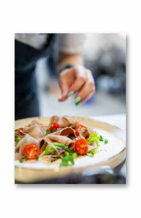 A close-up of a chef’s hands garnishing a gourmet dish with fresh green herbs. The plate features sliced meat, cherry tomatoes, and fresh greens, presented on a white plate