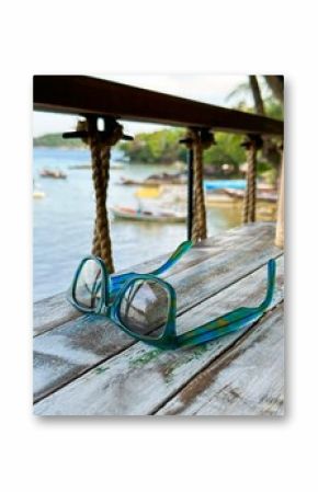 A close-up of colorful sunglasses resting on a rustic wooden table with a picturesque view of a tropical bay in the background. The reflection in the lenses captures the serene landscape, including