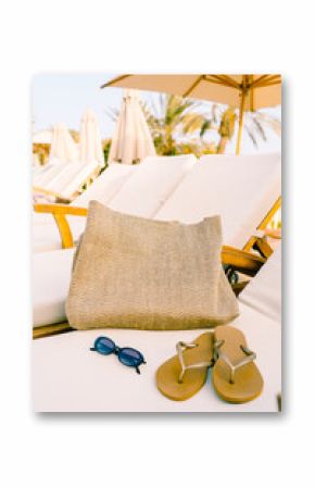 Close-up of a raffia beach bag, flip-flops, and sunglasses on a sun lounger under a parasol. Natural textures and warm tones evoke a relaxed, sustainable summer lifestyle.