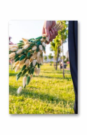 Elegant female hand holding soft fluffy bouquet of dried flowers while sitting on green grass in a black pleated dress. Summer evening outdoor scene.