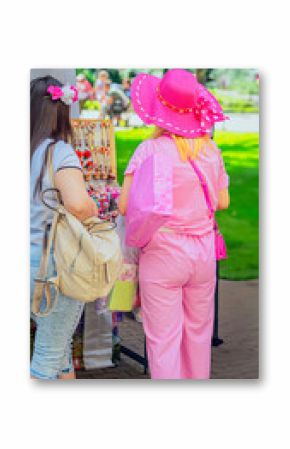 Two women explore outdoor market stand filled with handmade rose accessories and floral headbands on a sunny day. Traditional festive rose festival in Bulgaria, festival shopping