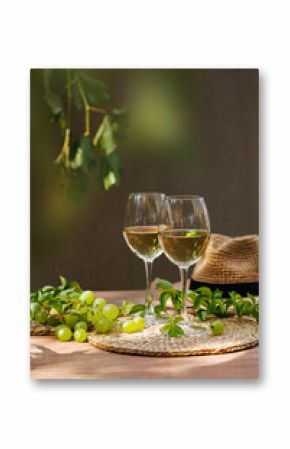 Two white wine glasses surrounded by grapes, vines and straw hat on sunny wooden table..