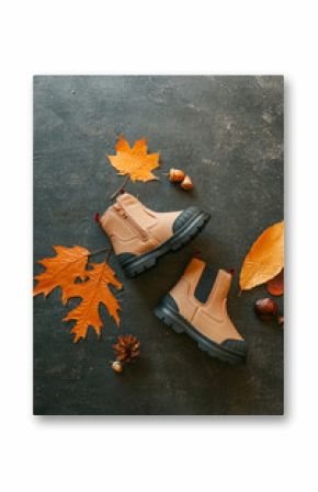 Children's beige boots with black soles on a dark textured background with autumn leaves, acorns, and pinecones. Stylish fall footwear for kids