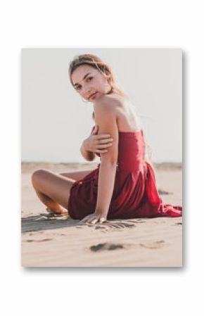 Attractive young lady with long blond hair wearing stylish red dress sitting on coast while looking at camera