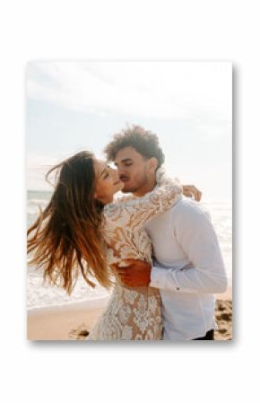 Side view of cheerful young bride and groom in stylish wedding costumes standing embracing each other with eyes closed on sandy beach near waving sea and enjoying each other