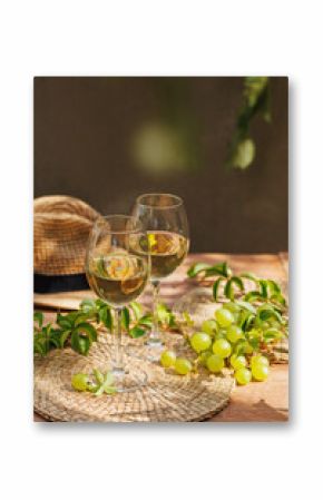 White wine glasses and bunch of grapes on rustic table with green vines and straw summer hat..