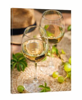 Two glasses of white wine with green grapes and straw hat in bright outdoor setting..