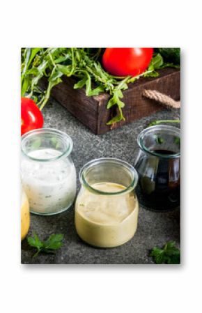 Set of dressings for salad: sauce vinaigrette, mustard, mayonnaise or ranch, balsamic or soy, basil with yogurt. Dark stone table. On background of greenery, vegetables for salad. Copy space