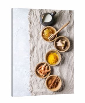 Ingredients for turmeric latte. Ground turmeric, curcuma root, cinnamon, ginger, honeycombs in wooden bowls, jug of milk over grey texture background with textile linen. Top view, copy space