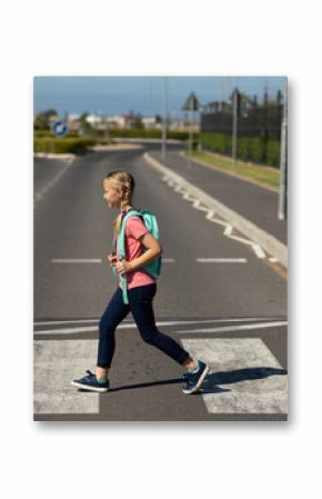 School-age girl crossing two-lane road on zebra crosswalk near shrubs, carrying mint green backpack