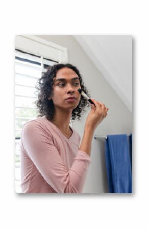 Non-binary person applying makeup brush to cheek in bathroom near white shutters and blue towel