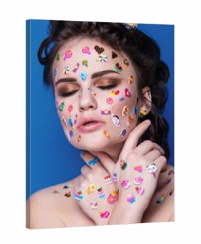 Beautiful fashion girl with luxury professional makeup and funny emoji stickers glued on the face. Young woman with cupcake in hands.
