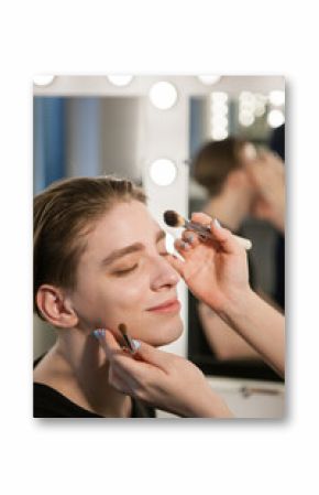 Men's makeup. A happy man will enjoy the make-up process. Make-up artist puts eye shadow and powder on face with brushes. Vertical indoors shot.