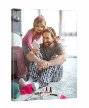 You are pretty fairy. Satisfied girl is showing mirror to her daddy after doing make-up. Man is looking at reflection with joy and laughing while sitting on floor in apartment 