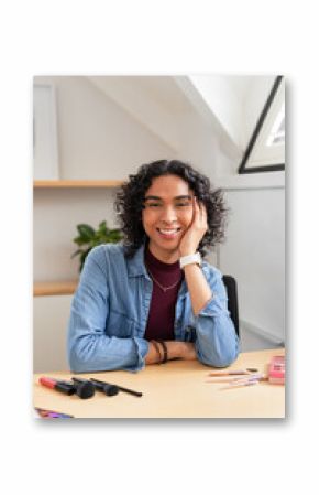 Non-binary person smiling while resting head on hand at attic desk with makeup palette and brushes