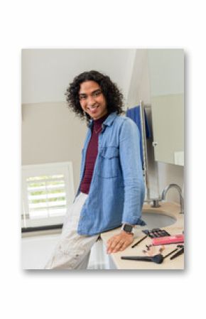 Non-binary adult leaning on bathroom counter by modern sink, smiling while arranging makeup brushes