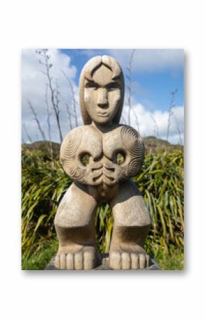 A powerful Maori carving, representing cultural heritage, stands among native New Zealand flax plants under a blue sky. Mercer Bay, Karekare, Auckland, New Zealand. 14 September 2024