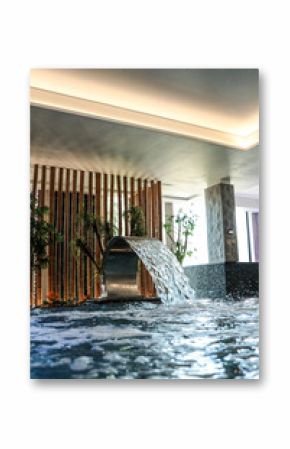Modern indoor spa pool with natural lighting, tropical plants, and a waterfall feature. Stylish relaxing zone with stone pillars and ceiling lights, evoking tranquility and luxury