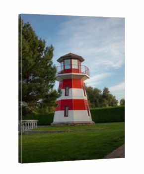 Red and white striped lighthouse on a green lawn, surrounded by trees and hedges under a blue sky, scenic coastal architecture and peaceful outdoor landscape view.
