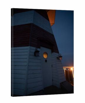 Close-up of a red and white striped lighthouse door at night, with warm light glowing through a round window and wall lanterns illuminating the exterior.