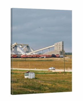 A potash plant with dome storage buildings, conveyor system, and trains across farmland with a dusty road under cloudy skies in Saskatchewan, Canada.