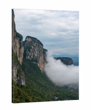 Jagged mountains rise steeply from a green valley, with clouds flowing around their peaks in Enshi City, China, showing majestic natural beauty.