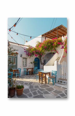 A picturesque alley in Paros features a traditional house surrounded by bright bougainvillea. Outdoor tables create a welcoming atmosphere for visitors to relax and enjoy the view.