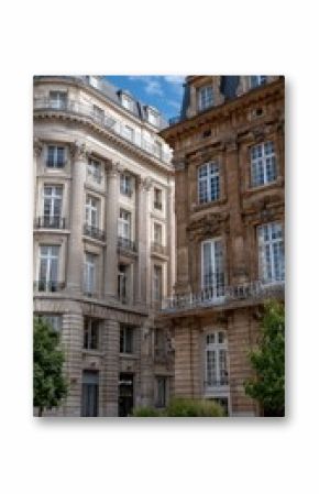 Beige and light brown buildings display large windows and elegant balconies, while trees line the street beside a charming boutique store
