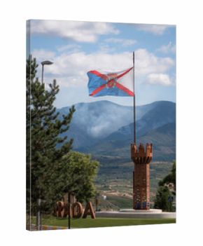 A flag of El Bierzo flies from a flagpole on a decorative tower while a mountainside burns in the background, causing large curtains of smoke during the devastating wildfires of 2025