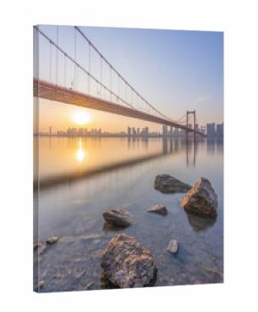 A long bridge spans a calm river at sunrise, with rocky shores in the foreground and a city skyline in the distance.