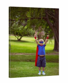 Boy standing on grassy park lawn, raising arms, wearing red cape, blue mask, red wristbands, bench