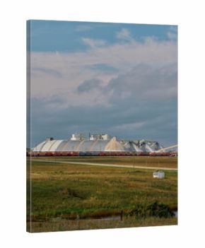 A potash plant with dome-shaped storage buildings, conveyor system, and train cars across farmland under cloudy skies in Saskatchewan, Canada.