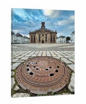 Blick auf einen Kanaldeckel mit Saarbrücker-Wappen und Schriftzug - im Hintergrund die  Ludwigskirche und der Ludwigsplatz