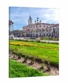 The pink-hued, clock-towered Alcalá City Hall provides a backdrop to the Plaza de Cervantes's formal gardens, featuring rows of yellow flowers, green grass, and pollarded plane trees