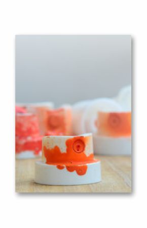Several plastic nozzles from a paint sprayer that lie on a wooden surface against a gray wall background. The caps are smeared in orange paint. The concept of street art and graffiti