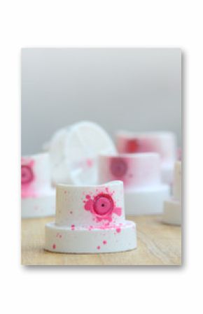Several plastic nozzles from a paint sprayer that lie on a wooden surface against a gray wall background. The caps are smeared in pink paint. The concept of street art and graffiti
