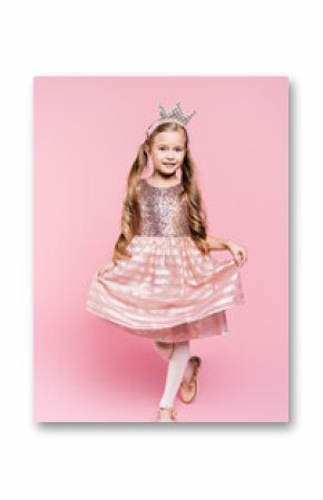 full length of cheerful little girl in dress and crown posing on pink