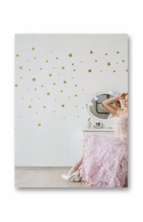 Attractive blond girl in princess pink knitted dress sitting near the makeup table and mirror with golden stars on white wall background