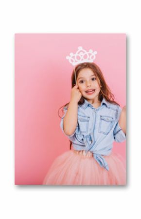 Joyful little girl with long brunette hair in tulle skirt holding princess crown on head isolated on pink background. Celebrating brightful carnival for kids, expressing positivity of birthday party