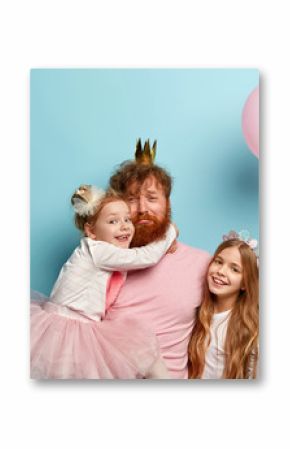 Tired sad single father organizes real holiday for children, wears crown, receives hug from small daughter, little girl with long hair holds air balloon, smiles happily stands near. Family celebration