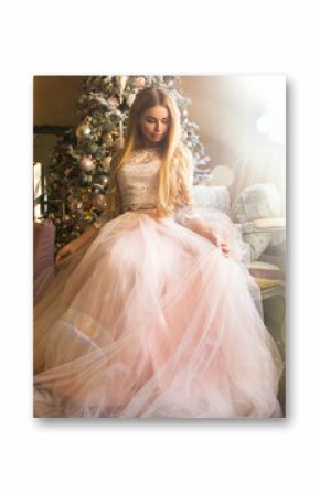 young happy blonde girl with long hair in pink princess dress is sitting near window, christmas tree and sofa in light interior. luxury honey woman with cute smile. chistmas concept, free space