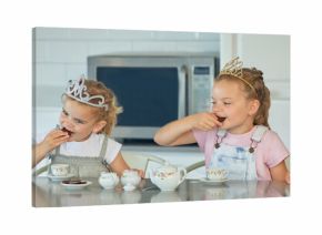 Two little girls having a princess tea party at home. Sibling sister friends wearing tiaras while playing with tea set and having cookies at kitchen table. Sisters getting along and playing together