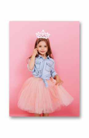 Cheerful little girl with long brunette hair in tulle skirt holding princess crown on head  isolated on pink background. Celebrating brightful carnival for kids, birthday party, having fun of cute kid