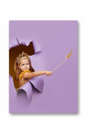 Cute little child girl in princess costume breaks through a colored purple paper wall. Points with a magic wand to the empty space on the right. Toddler funny emotions face.