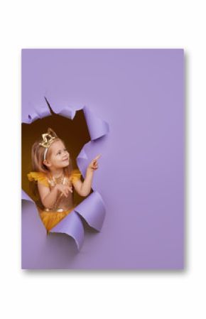 Cute little child girl in princess costume breaks through a colored purple paper wall. Points with a magic wand to the empty space on the right. Toddler funny emotions face.